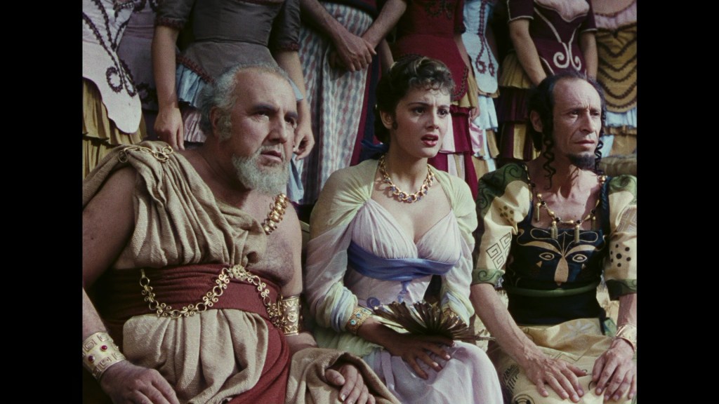 Three characters in ancient attire express concern, seated in an audience. The man on the left has a beard and wears a toga, the woman in the middle has curly hair and a dress with draped fabric, and the man on the right has a long hairstyle adorned with decorative motifs.