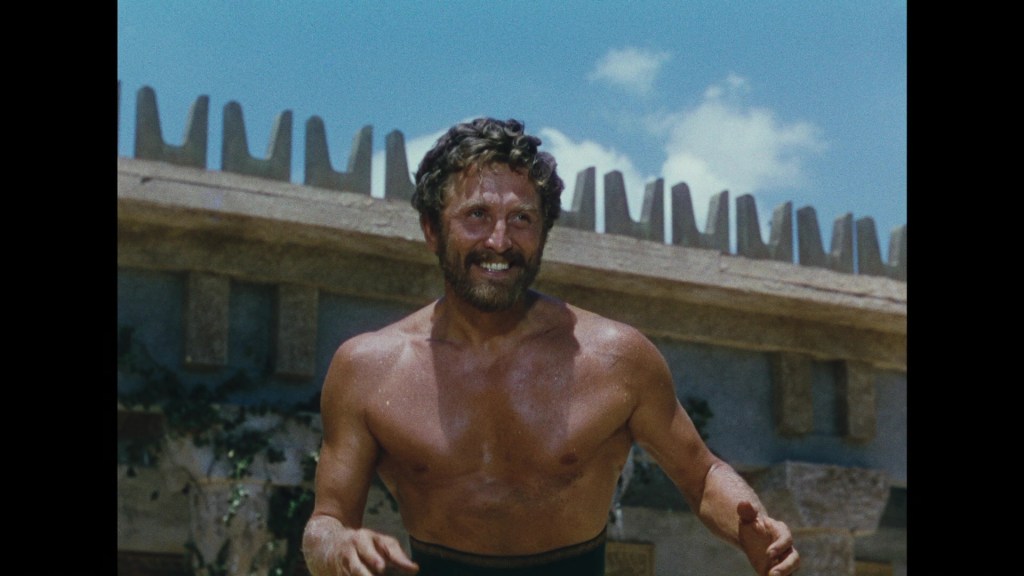 A smiling man with a beard and curly hair, wearing no shirt, stands outside against a blue sky and ancient ruins backdrop.