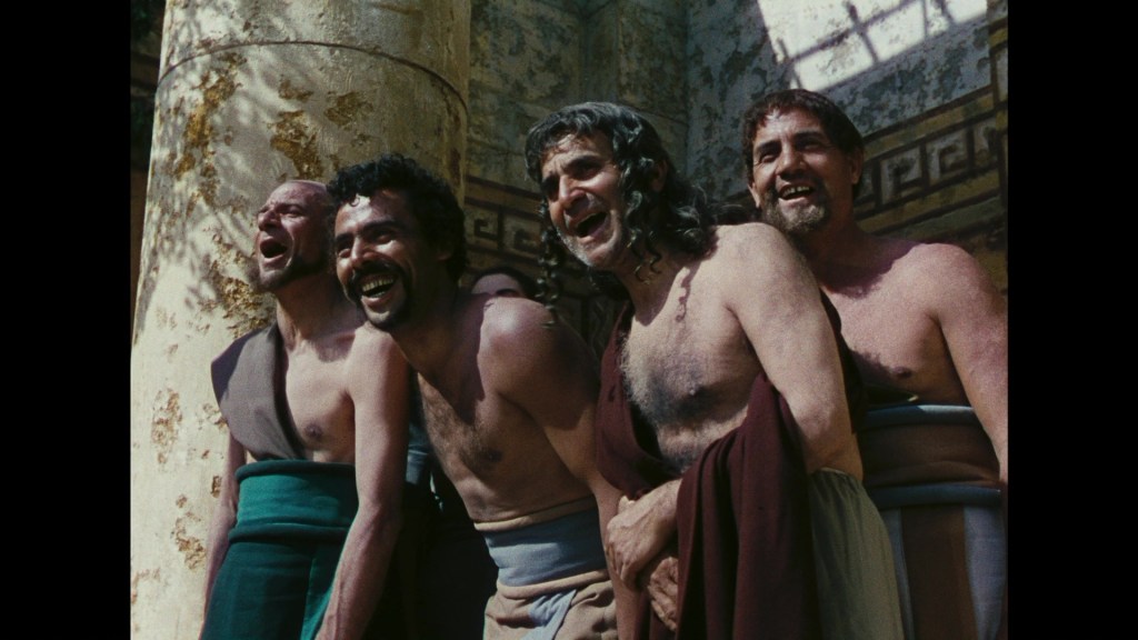 Group of five men smiling and laughing, wearing ancient tunics, standing in front of a stone wall with classical Greek architecture details.