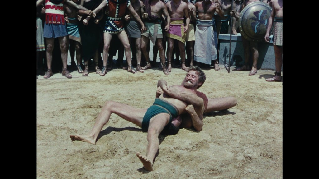 A wrestling match taking place in an ancient setting, featuring two male wrestlers on the ground, surrounded by an enthusiastic crowd watching the competition.