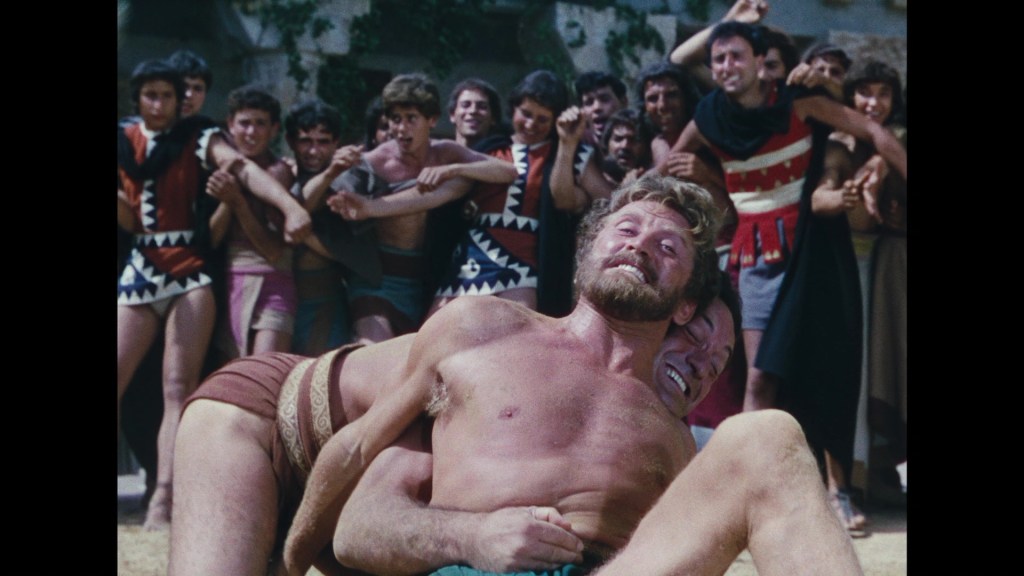 Two men wrestle in an ancient arena, while a cheering crowd in colorful attire watches the match.