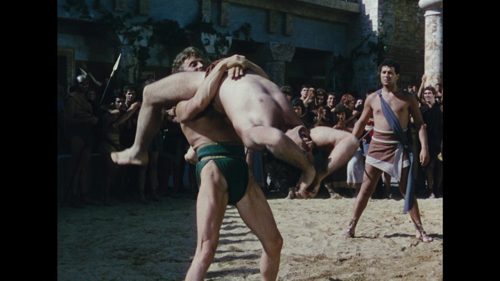 Two wrestlers compete in a sandy arena, with one lifting the other in a display of strength, while an audience watches in the background.