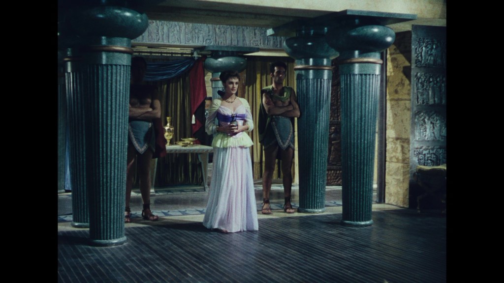 A woman in ancient Roman attire stands between two tall columns, with two men in tunics standing behind her in a decorated interior setting.