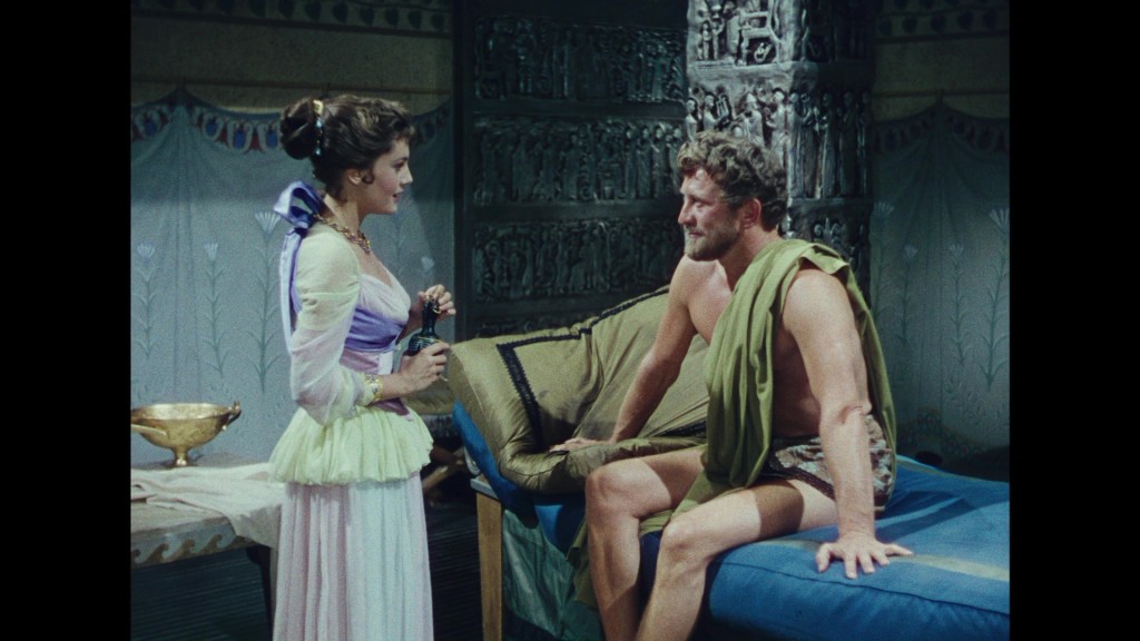 A woman in a historical dress talks to a man wearing a toga, seated on a bed with a decorative backdrop.