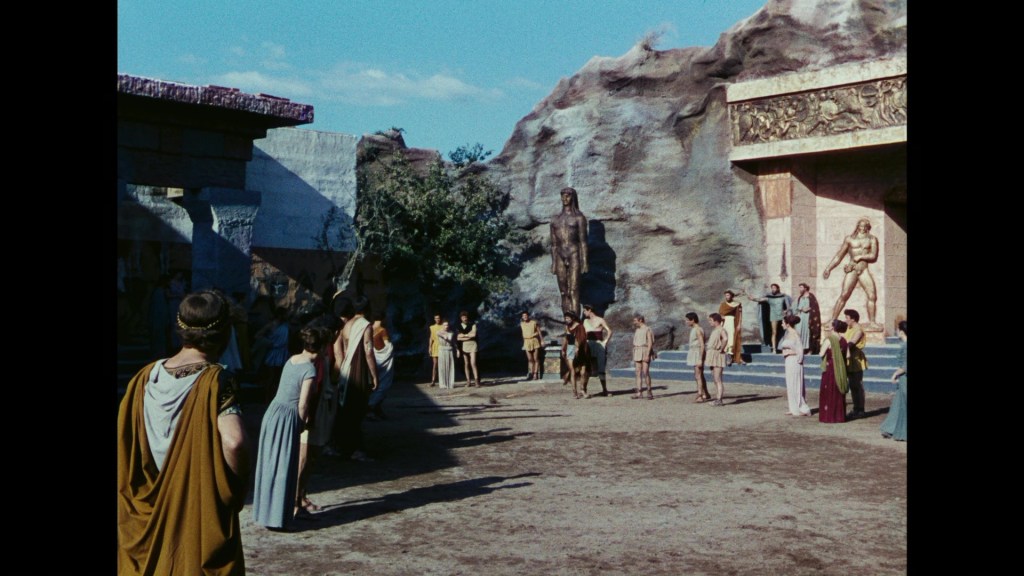 A scene depicting ancient Greek-style figures in a courtyard surrounded by large statues and building structures, with individuals dressed in classical togas and attire engaging in a gathering or event.