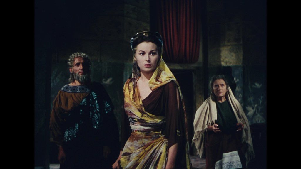A scene from a dramatic film featuring three characters in historical costumes: a man with curly hair, a woman in a colorful dress with a yellow shawl, and an older woman in subdued clothing. The background shows a dimly lit room with decorative elements.