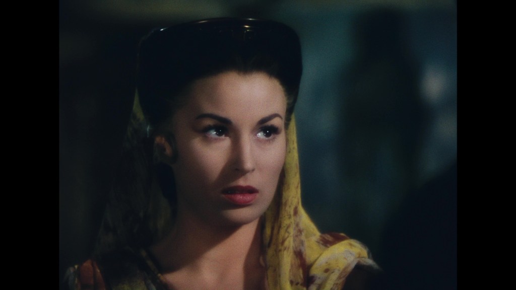 Close-up of a woman with dark hair and a yellow scarf draped over her shoulders, looking with a serious expression.