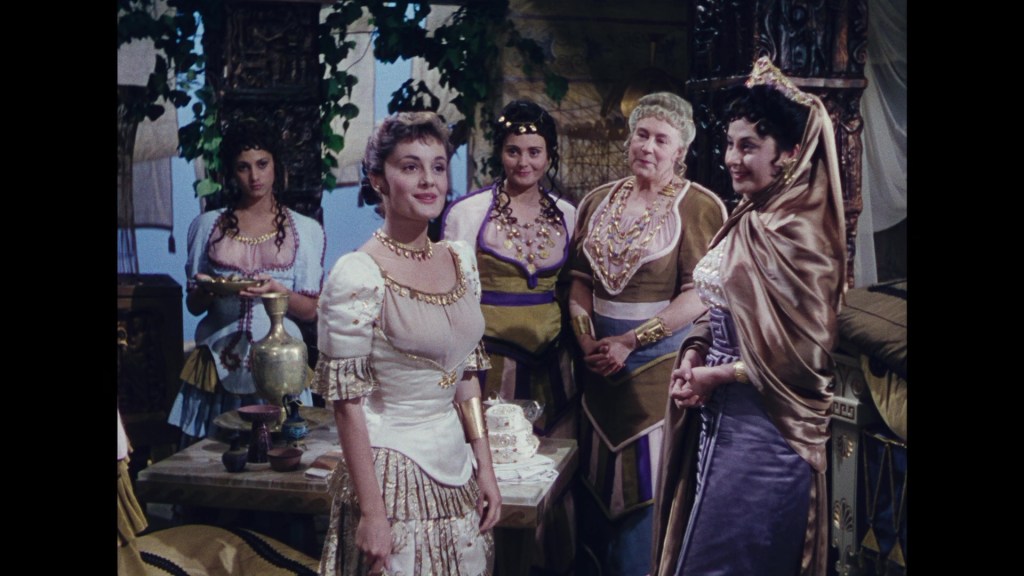 A group of five women dressed in elaborate, historical costumes, gathered in an ancient-themed setting, with greenery and decorative items visible in the background.