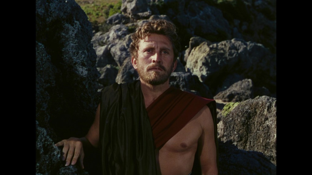 A man with curly hair, wearing a black garment draped over one shoulder, stands among large rocks, looking thoughtfully into the distance.