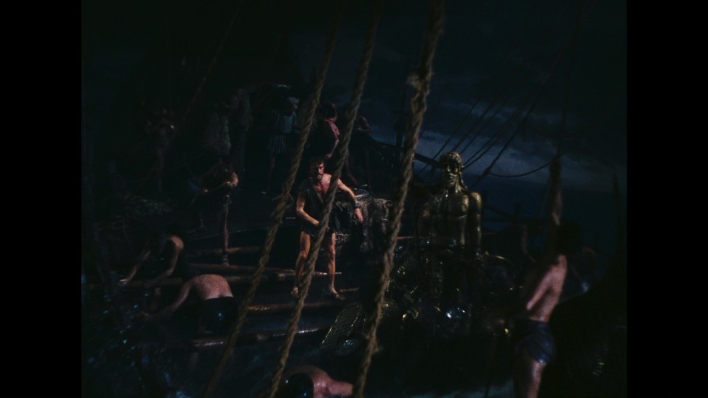 A scene from a film featuring sailors on a ship, with some working on ropes and others interacting with a large golden statue. The atmosphere is dark and stormy.
