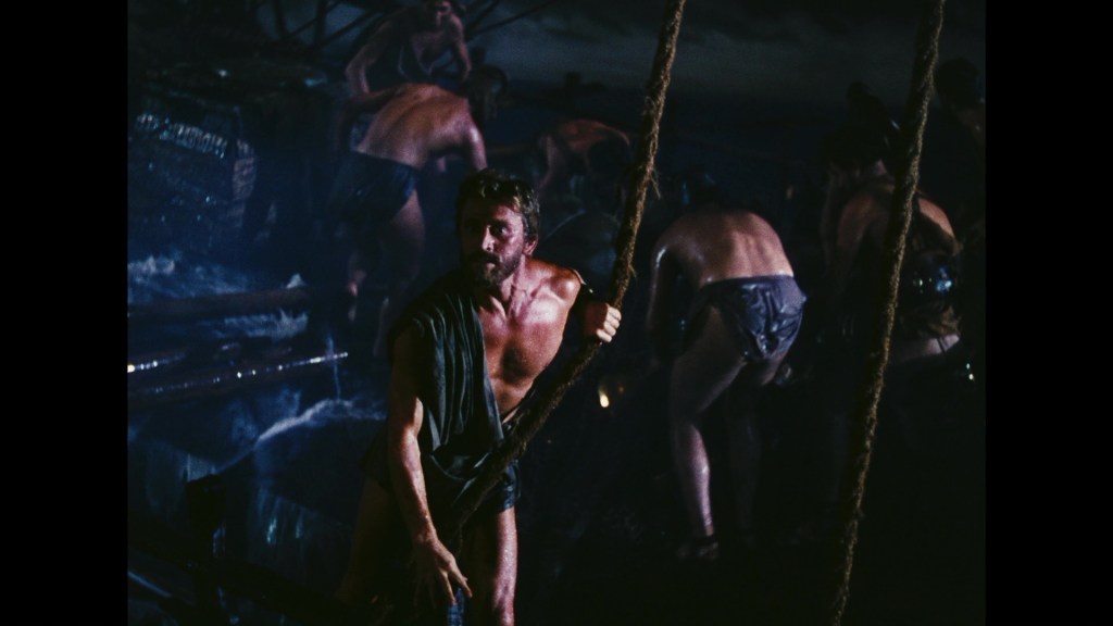 A group of men working on a ship during a tumultuous sea scene, with one man prominently pulling on a rope in the foreground.