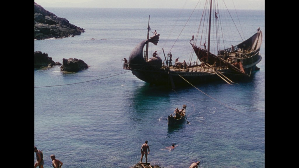 A scenic view of a coastline featuring a large, ornate ship resembling a whale, with several people on board and in the water, near rocky shores and a clear blue sea.