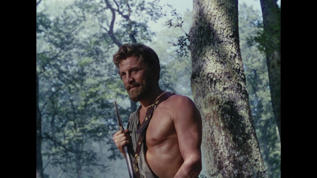 A muscular man with a beard stands in a forest, holding a spear. He is shirtless and appears to be in a thoughtful pose, surrounded by trees and dappled sunlight.