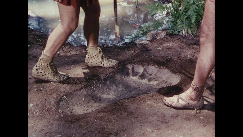 Two people in sandals stand near a large footprint in the ground, surrounded by water and greenery.