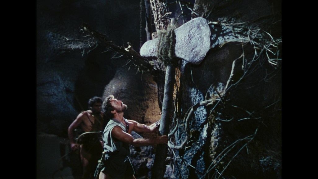 A prehistoric man lifting a large stone tool or weapon in a cave, with another figure in the background observing.
