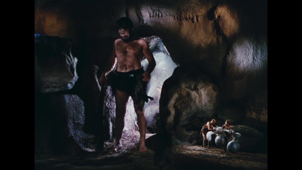 A muscular man stands in a rocky cave, looking down at two smaller figures who appear to be preparing large stone objects.