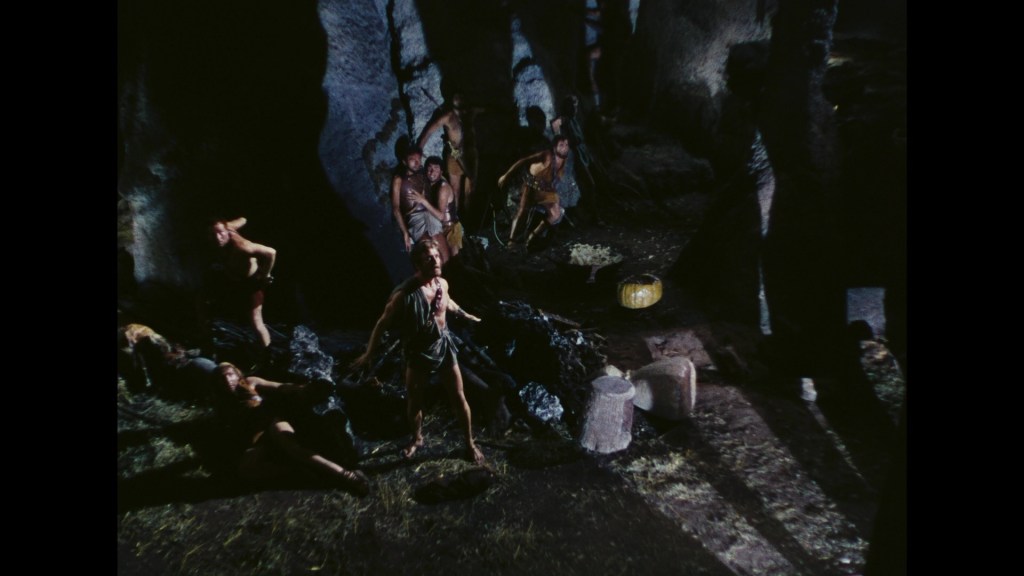 A group of individuals in primitive clothing inside a dark cave, with some appearing to be agitated or in motion.