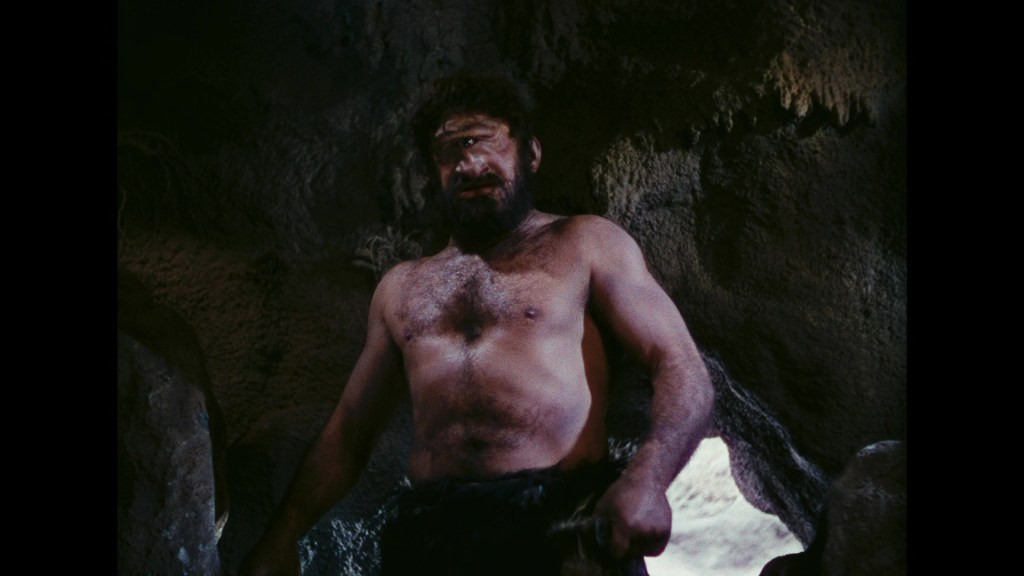 A muscular man with a beard, standing shirtless in a rocky cave setting, with a serious expression.