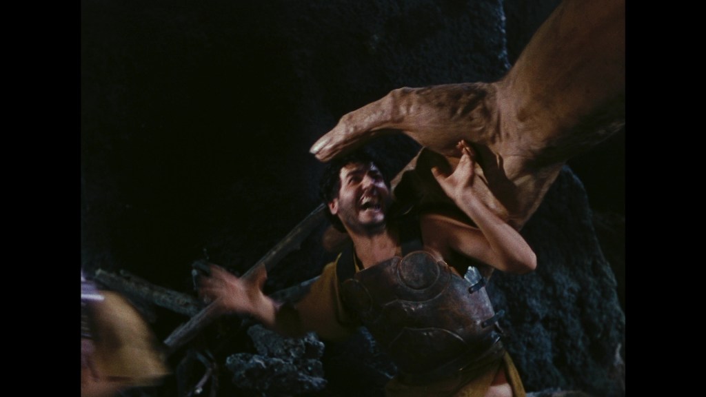 A man in a tunic and armor is depicted in a dramatic scene, appearing to be attacked or overwhelmed by a large creature's hand in a dark, rocky environment.