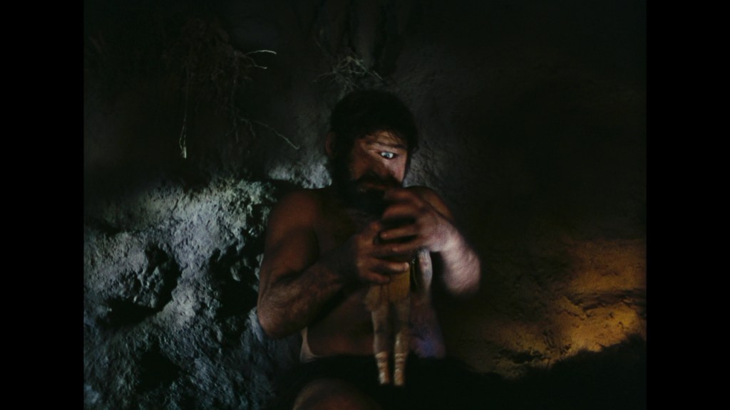 A man with a muscular build sits in a dimly lit cave, holding a small, doll-like figure in his hands, with a focused expression on his face.
