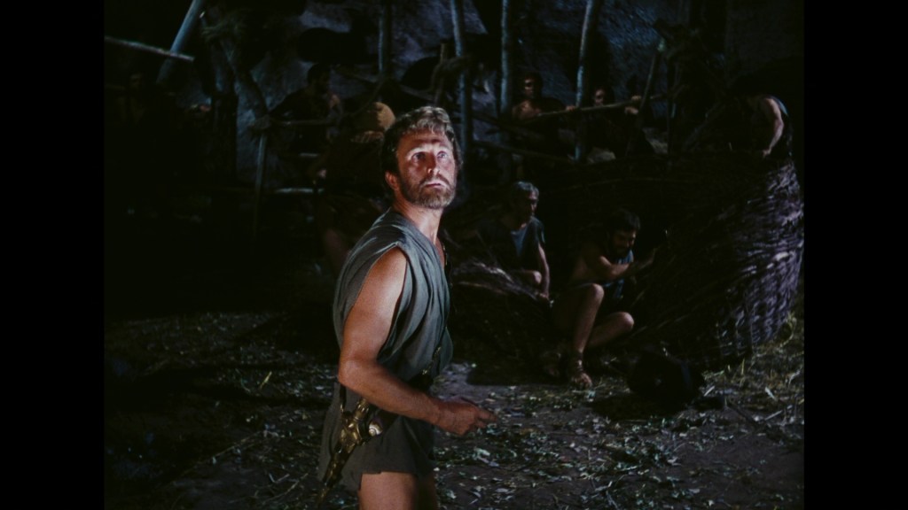 A tense scene featuring a man with a beard, wearing a tunic, looking upwards. He is surrounded by other figures in a dimly lit environment, suggesting an ancient or mythical setting.