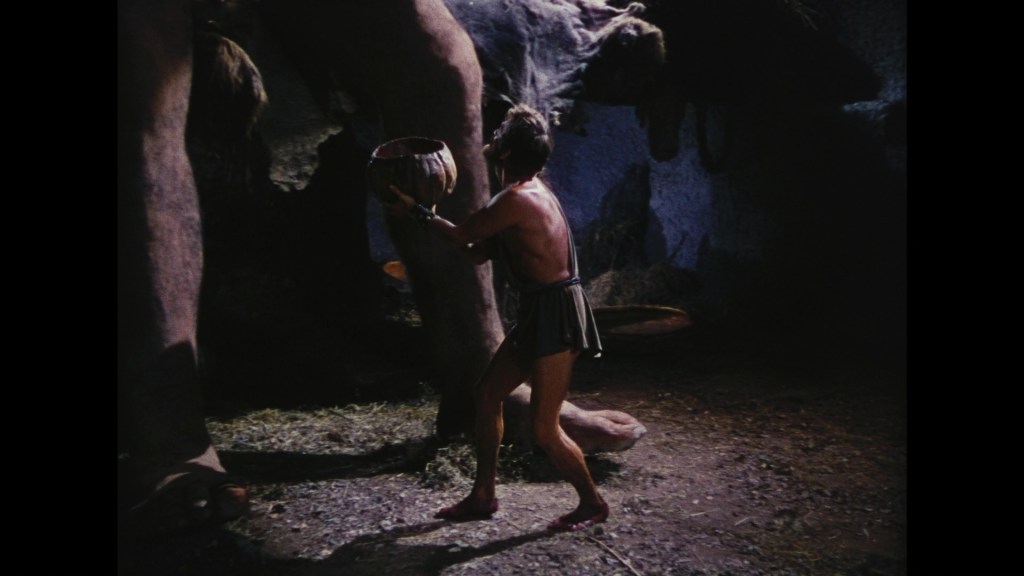 A man in a classic tunic uses a bowl while standing near a large, mysterious creature in a dimly lit cave.