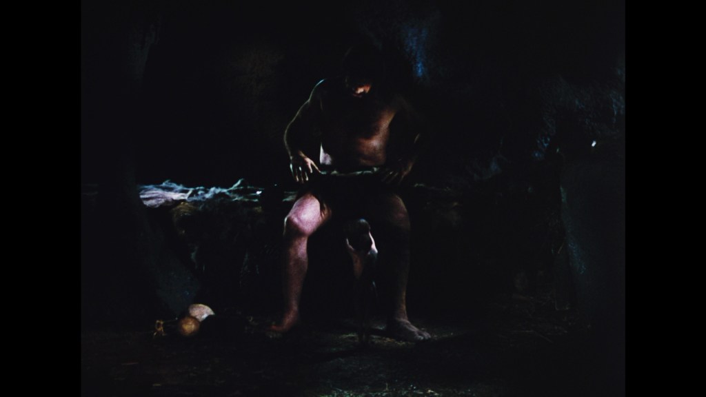 A dimly lit cave scene featuring a shirtless man sitting on a rock, appearing to craft or work on an object. Nearby, there are small items on the ground, such as gourds or potatoes.