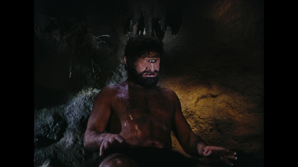 A muscular, shirtless man with a beard and short hair, sitting in a dark, cave-like environment with a surprised expression on his face.