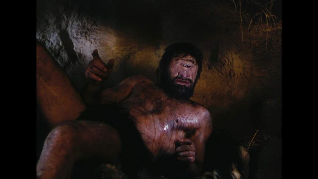 A muscular, bearded man with a single eye sits in a cave, gesturing with his right hand, showcasing a rugged environment.