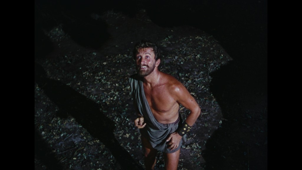 A muscular man in ancient attire gazes upward with a smile, surrounded by a dark forest floor covered in leaves.