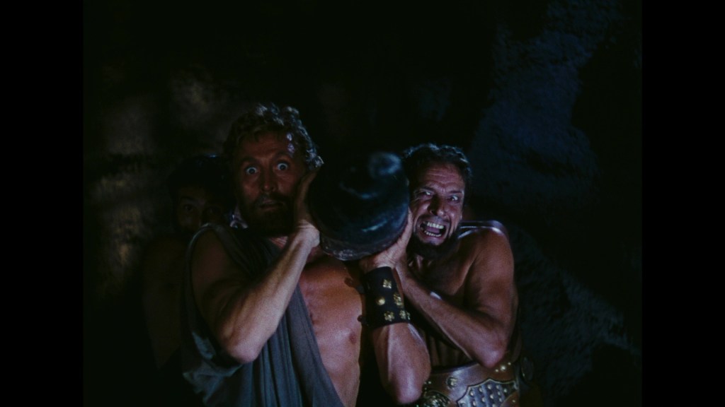 Two muscular men struggle to lift a heavy object while a third figure watches from the shadows, in a dark, cave-like setting.