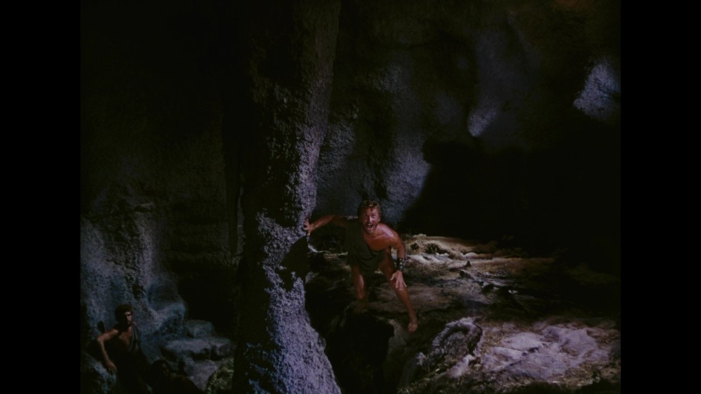 A dramatic scene set in a dark cave, featuring two muscular men, one standing against a rock and appearing startled, while the other is seen in the background.