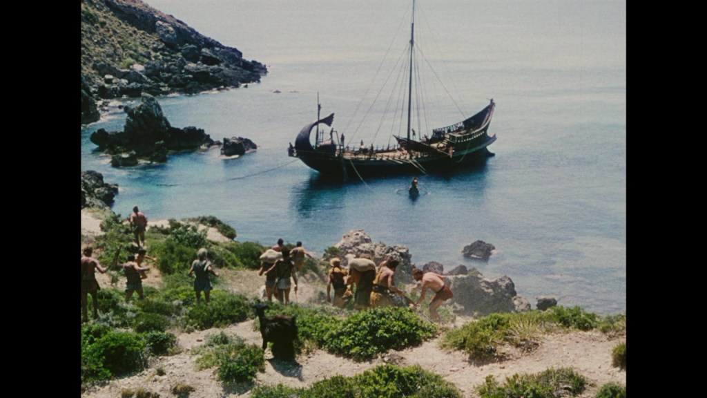 A group of ancient warriors is gathering on a rocky beach near a tranquil blue sea, with a large, traditional ship anchored offshore.