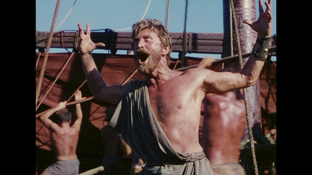 A muscular man with long hair and a beard yells with intense emotion on a ship, surrounded by other men working in the background.