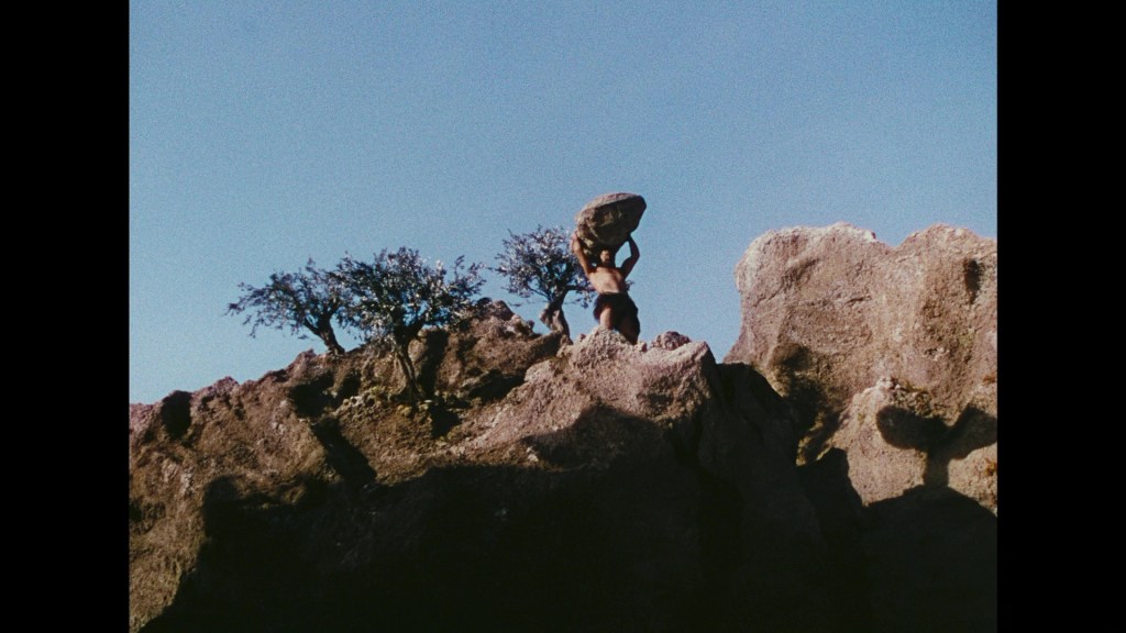 A person balances a large rock above their head while standing on a rugged mountain terrain, with small trees in the background against a clear blue sky.