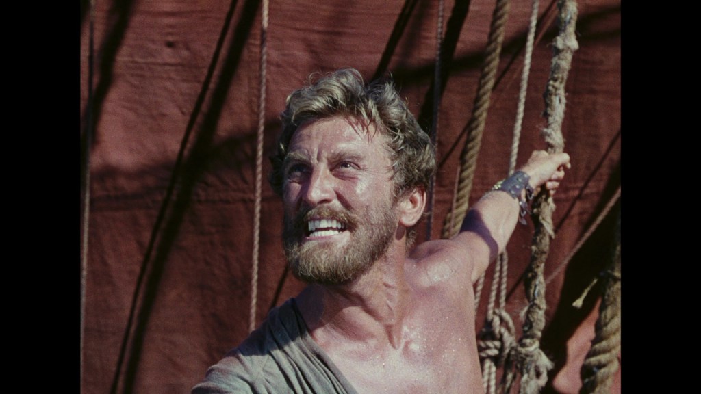 A muscular man with a beard, wearing a sleeveless top, is smiling and gesturing while standing near ropes on a ship.