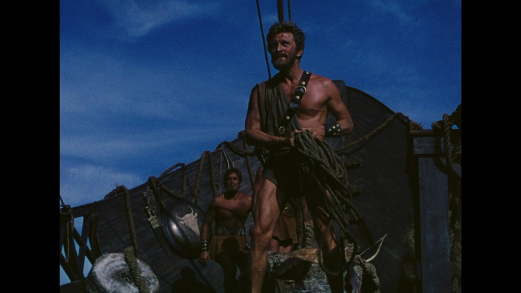 A muscular man in ancient Greek attire stands on a ship deck, holding ropes, with blue sky in the background. Another man is visible in the background.