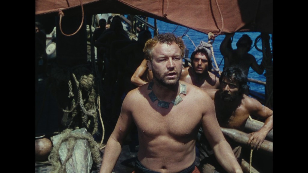A shirtless man with a necklace stands on a ship surrounded by crew members in the background, with a blue ocean visible.
