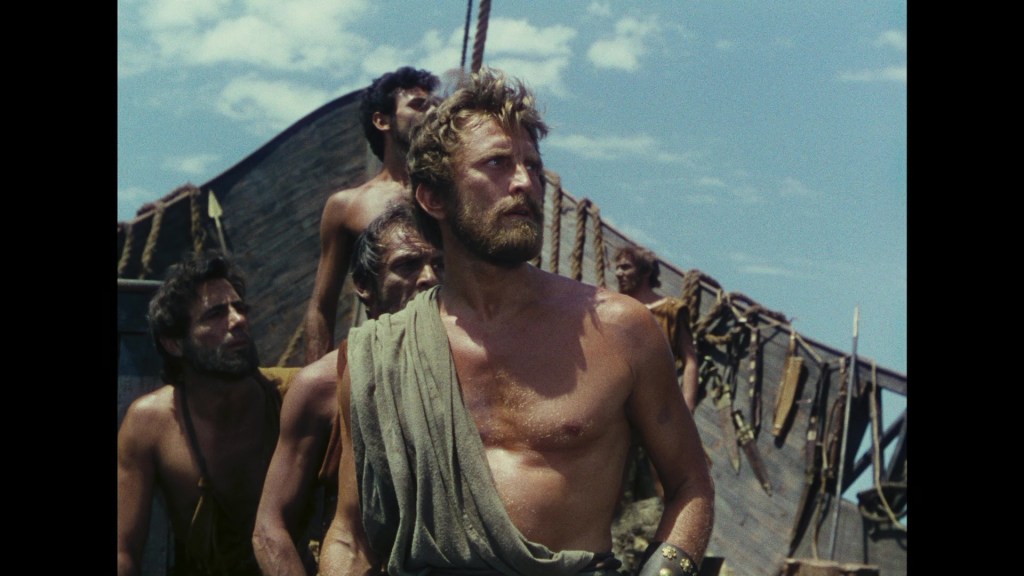 A group of muscular men aboard a ship, with one man in the foreground wearing a tattered cloth and looking intently into the distance against a bright sky.