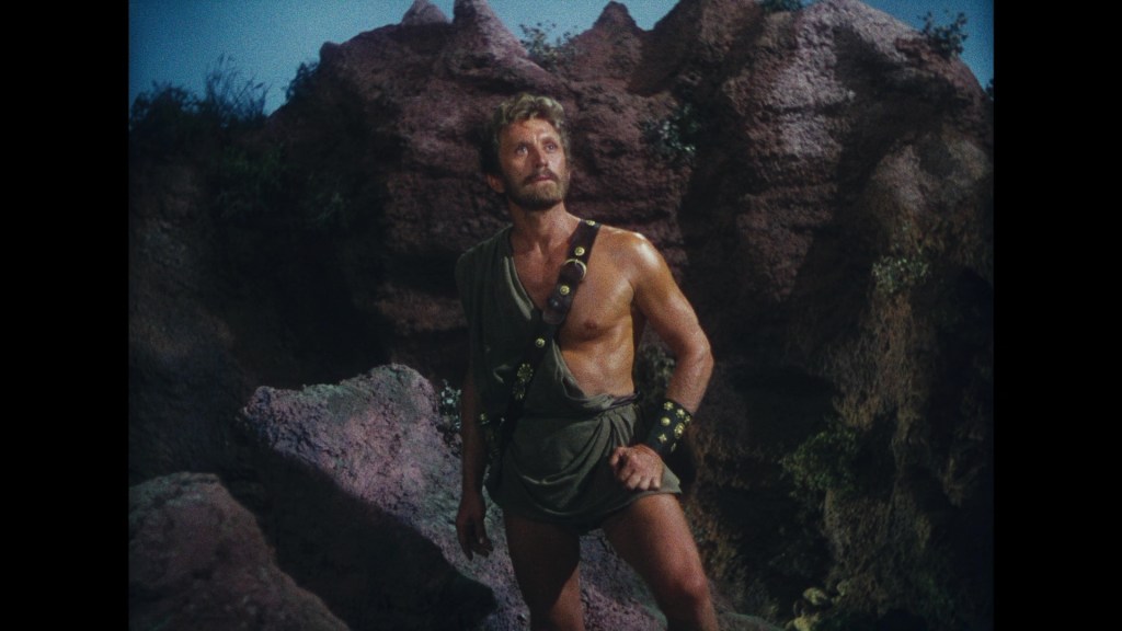 A muscular man wearing a tunic stands on rocky terrain, looking upward with a thoughtful expression.