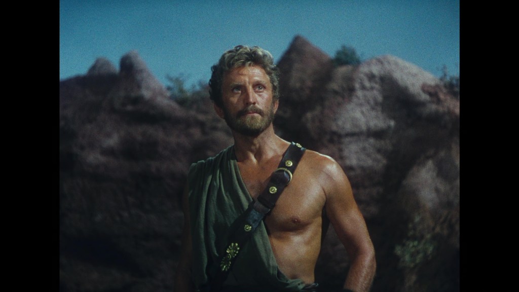 A muscular man with a beard, dressed in a tunic, stands in front of rocky terrain under a blue sky, looking confidently into the distance.