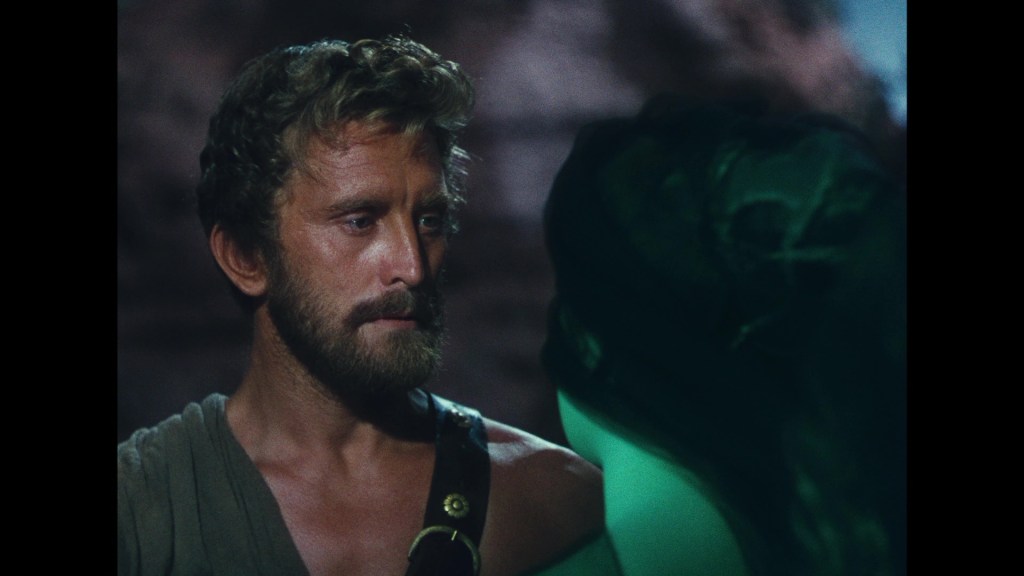 Close-up of a man with curly hair and a beard, looking intently at a woman with dark hair, partially obscured in shadow, set against a rocky background.
