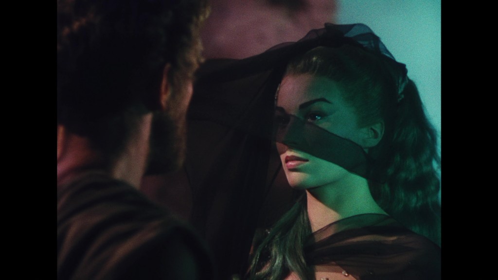 A dramatic scene featuring a man with a beard facing a woman draped in a sheer black fabric, highlighting her facial features and long hair, illuminated with colorful lighting.