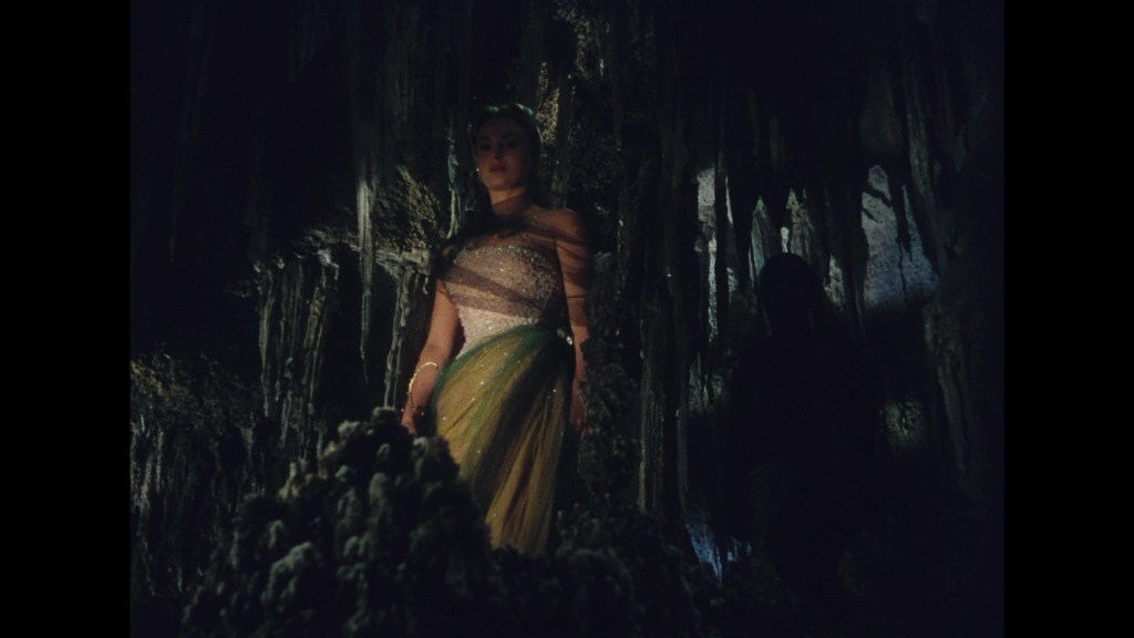 A woman in a flowing gown stands in a dark cave, surrounded by stalactites and rock formations, with shadows in the background.