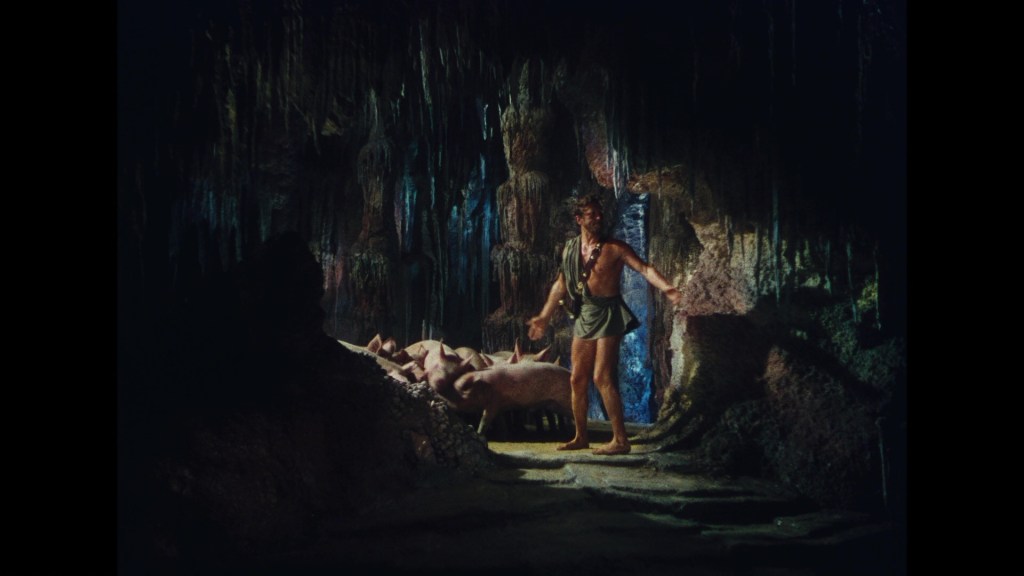 A man in minimal attire stands inside a cave, surrounded by stalactites and dim lighting, with several pigs nearby.