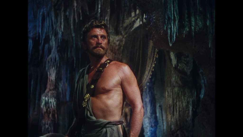 A muscular man with a beard, wearing a toga, stands in a cave-like environment with textured rock formations and hanging stalactites.