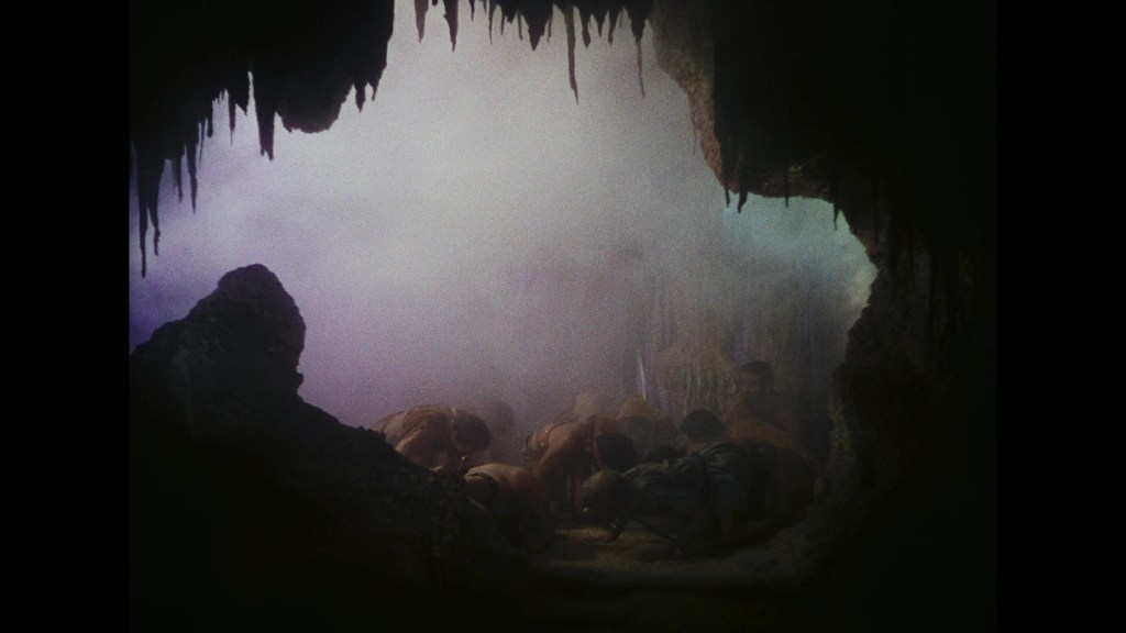 A dimly lit cave interior showing several figures huddled on the ground, surrounded by smoke and rock formations.