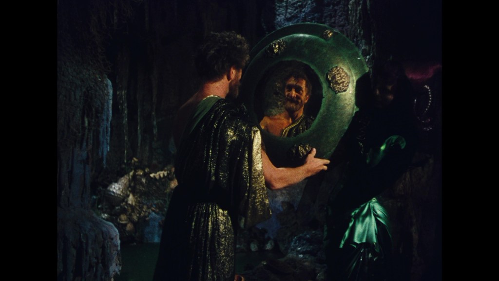 A man in a shimmering green robe gazes into a circular mirror held by another figure, reflecting a bearded man with an ornate background in a dark, cave-like setting.