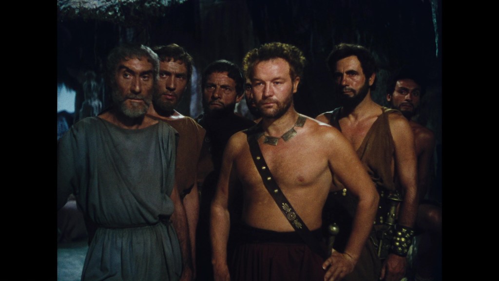 Group of men in ancient Greek clothing, standing together with serious expressions in a dimly lit setting.