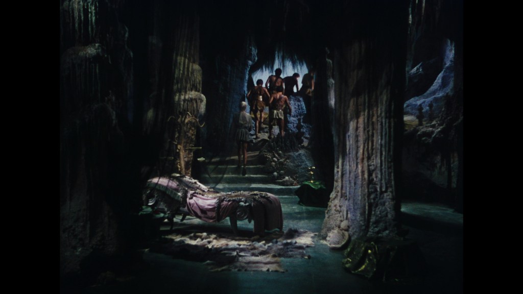 A dark, cave-like interior with rugged rock formations. A group of people stands in the background, while a bed draped with fabric is in the foreground.